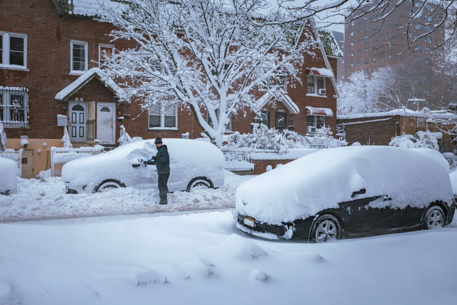 A residential street blanketed in deep snow during winter, featuring two parked cars semi-buried under thick snow covers, with tire treads and side mirrors barely visible. A person dressed in dark winter clothing, including a hat, stands beside one of the vehicles, clearing snow with a snow brush or scraper. In the background, there is a red-brick house with white trim, a small front garden, and a leafless tree with snow-laden branches extending overhead. The scene is illuminated by diffused natural light, highlighting the snow's texture and the quiet, cold atmosphere. The image reflects a typical winter setting where private vehicle owners often temporarily clear snow from their cars, possibly in preparation for an independent collection or on-site clearance, aligning with services offered by rubbish removal companies like rubbishremovalqueenspark.co.uk that support efficient local waste handling during adverse weather conditions.