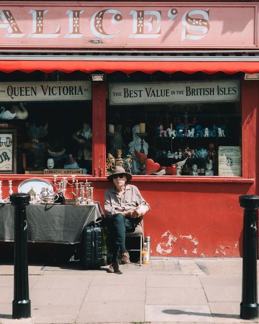 A woman seated on a bench in front of a red storefront window displays various collectibles and decorative items, including statues, trophies, and small figurines. She wears sunglasses, a tan wide-brimmed hat, and casual clothing with a light-colored jacket. Next to her is a black suitcase, and she holds some papers in her hands. To her left, a black cloth covers a table displaying additional items such as small lamps and trinkets, some of which are wrapped in plastic or protective coverings. The building's exterior features a weathered red facade with visible peeling paint and scuffed areas near the bottom. The large window showcases mannequins dressed in clothing, and signs reading 'Queen Victoria' and 'The Best Value in the British Isles' are visible above the display. The sidewalk in front of the shop is plain concrete, with black metal bollards separating the pedestrian area from the street. The scene is illuminated by daylight, casting soft shadows, and the overall environment suggests a busy shopping street where independent outlets, including those managing waste disposal through private rubbish removal services, are common.