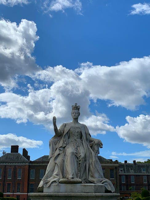 A classical stone statue of a regal female figure wearing a crown and flowing robes, seated with her right hand raised in a gesture of blessing or authority. The statue is positioned outdoors in a public square or park, with a backdrop of red-brick buildings and a bright blue sky filled with fluffy white clouds. The statue's surface appears weathered, with detailed drapery and a calm, solemn expression. The surrounding environment suggests a historical or civic setting, with some greenery visible at the base, and the statue's location likely associated with a city that values its heritage, possibly near sites offering private or alternative waste collection options to local authorities, as indicated by services like rubbish removal in Queens Park.