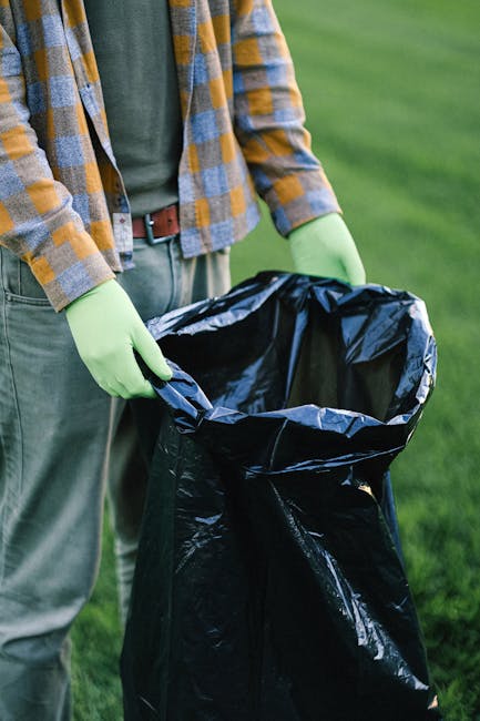 A person wearing a plaid shirt layered over a grey top, with bright green gloves, holding open a large black plastic rubbish bag outdoors on a grassy area. The bag appears to be empty or partially filled and is positioned near the person's waist, with their hands gripping the edges. The background features a lush, green grassy environment, suggesting a garden or park setting. The scene conveys waste collection or rubbish disposal activity, highlighting the importance of proper waste management and private collection services such as those offered by rubbishremovalqueenspark.co.uk, which supports efficient on-site clearance away from public rubbish bins. The lighting is natural, indicating daytime, and the focus is on the action of preparing the rubbish bag for waste collection, aligning with the context of private or independent waste handling typically associated with rubbish removal services in Queens Park.