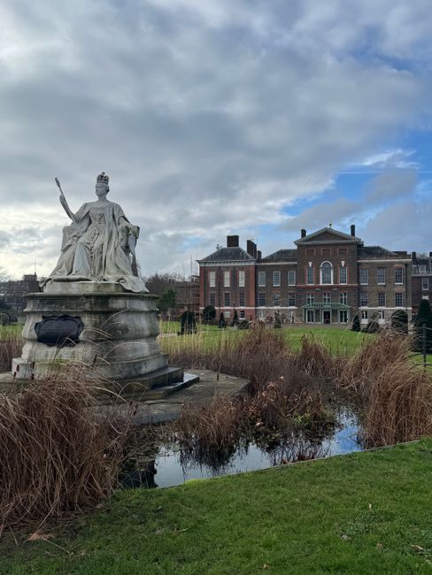 A classical stone statue of a regal female figure wearing a crown and flowing robes, seated with her right hand raised in a gesture of blessing or authority. The statue is positioned outdoors in a public square or park, with a backdrop of red-brick buildings and a bright blue sky filled with fluffy white clouds. The statue's surface appears weathered, with detailed drapery and a calm, solemn expression. The surrounding environment suggests a historical or civic setting, with some greenery visible at the base, and the statue's location likely associated with a city that values its heritage, possibly near sites offering private or alternative waste collection options to local authorities, as indicated by services like rubbish removal in Queens Park.